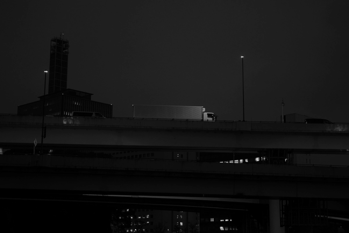 A truck crossing an elevated highway at night, in black and white, with a tower and port buildings behind.