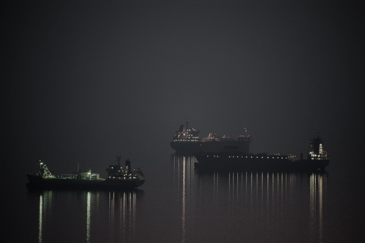 Three cargo ships waiting at anchor in fog at night, their lights reflected in still water.