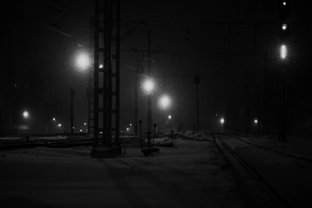 Rail tracks at night with snow falling through the light of trackside lamps.